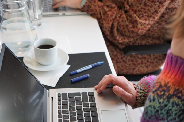 En person sidder ved en laptop på et skrivebord. Personens hånd hvilker på keyboardet. Ved siden af ligger kuglepenne og en kop kaffe.